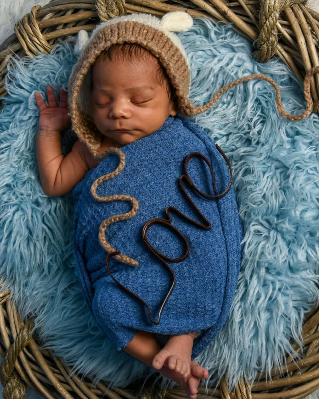 Newborn baby wrapped in a blue blanket with 'love' embroidery, wearing a brown knitted hat, lying on a textured blue surface.