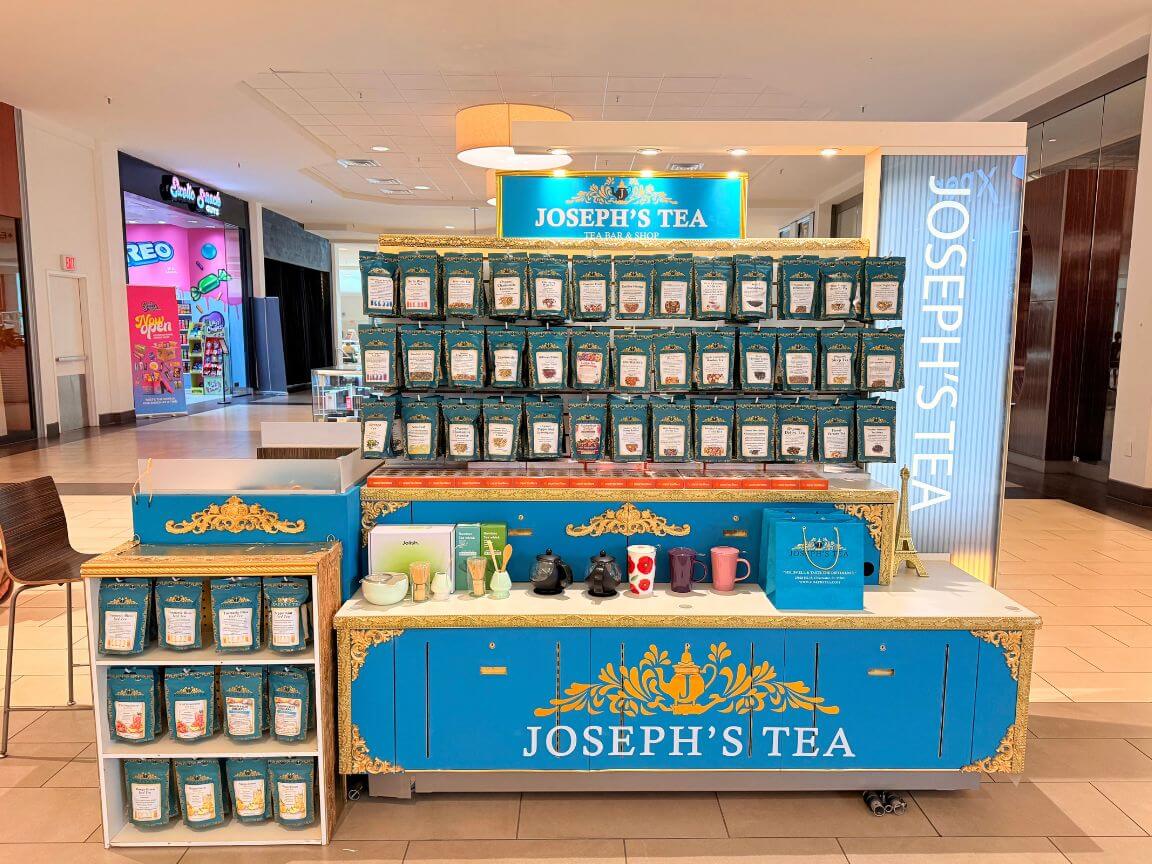 A Joseph's Tea branded display cart in a shopping mall, featuring multiple rows of packaged tea bags, a counter with teapots and mugs, and side shelves of additional tea packages.