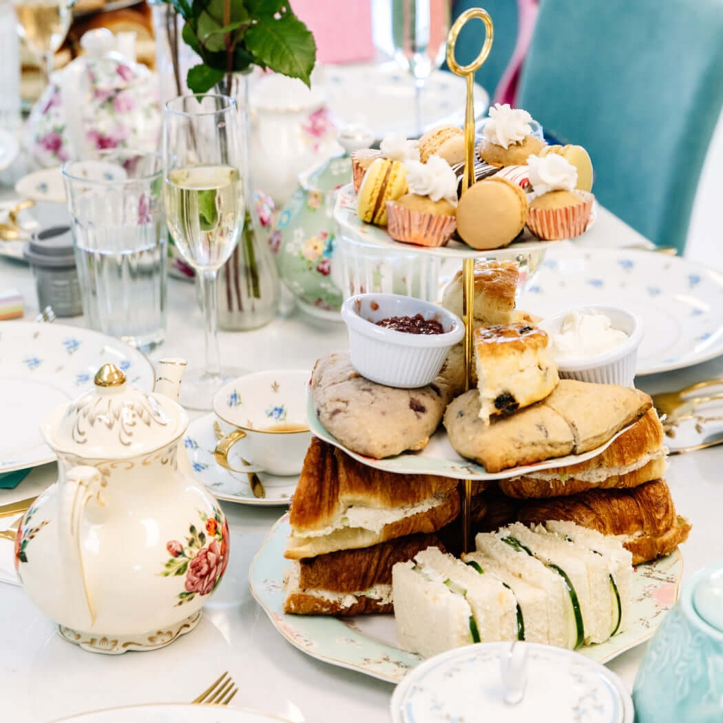 Three-tiered cake stand with assorted pastries on a table setting with tea pots and glasses.
