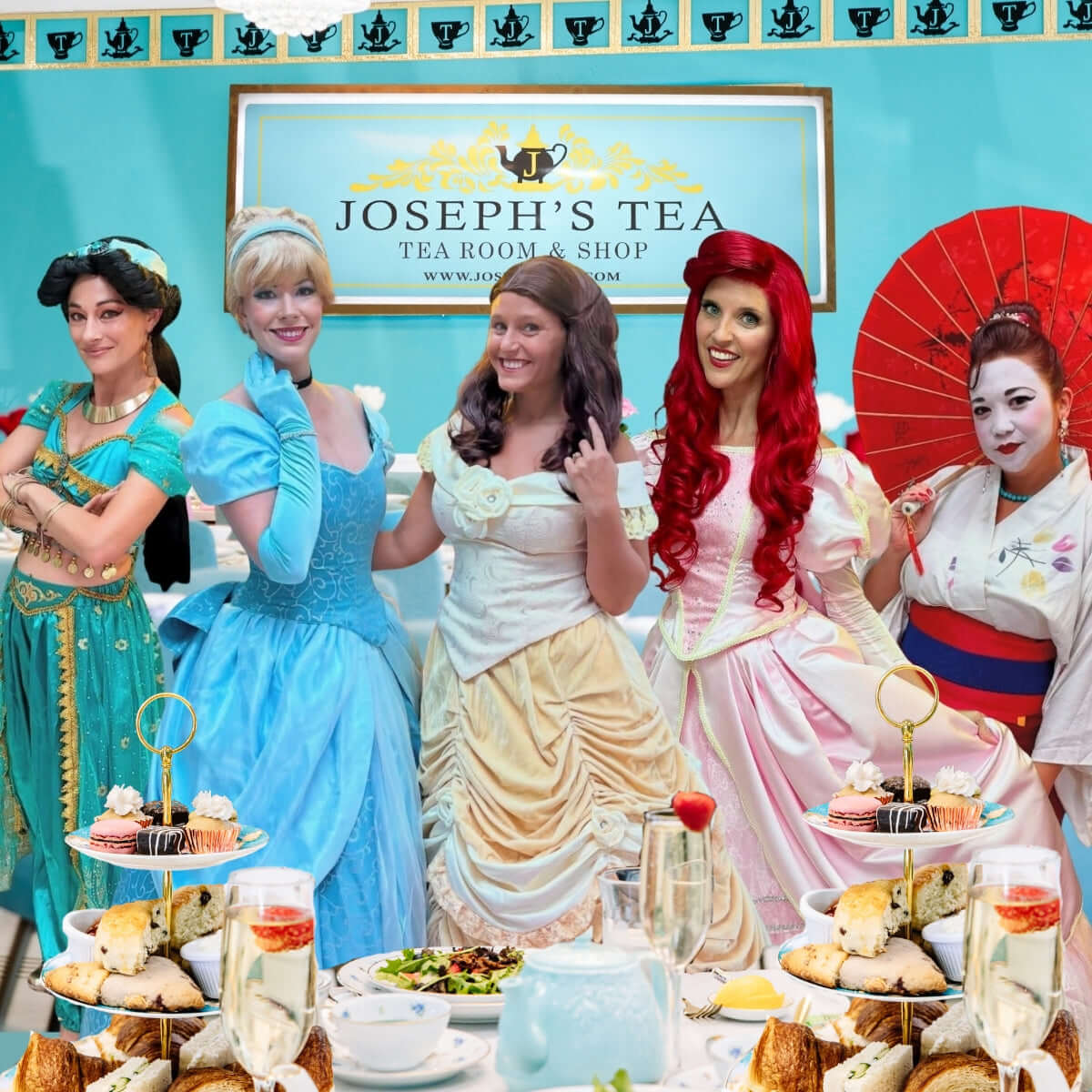 Five women in colorful costumes posing in front of a 'Joseph's Tea' sign with a tea party setup.