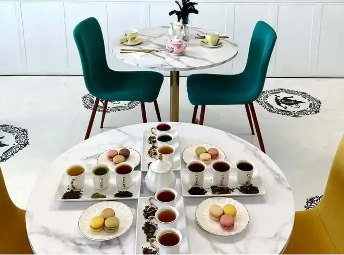 A styled cafe interior with two round white marble tables set with cups of tea, pastries, and macarons, surrounded by teal and yellow chairs against a turquoise wall with framed art.